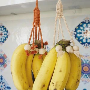 Puede incluir: Dos colgadores de pl&aacute;tanos de macram&eacute;, uno naranja y otro crema, cada uno con un racimo de pl&aacute;tanos amarillos maduros. Los colgadores est&aacute;n suspendidos de ganchos met&aacute;licos. El texto "Banana Hanger" est&aacute; en la parte superior de la imagen.