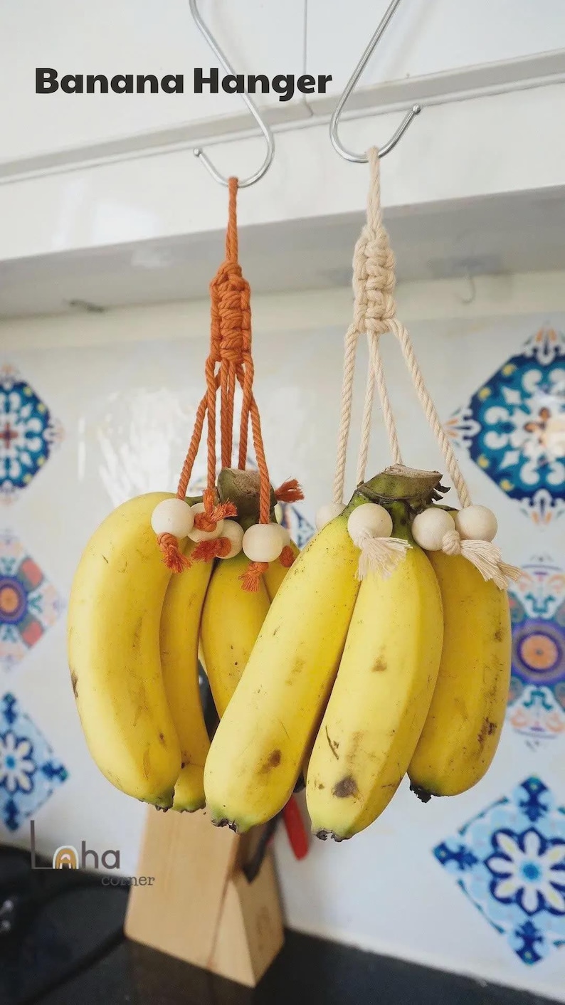 Puede incluir: Dos colgadores de pl&aacute;tanos de macram&eacute;, uno naranja y otro crema, cada uno con un racimo de pl&aacute;tanos amarillos maduros. Los colgadores est&aacute;n suspendidos de ganchos met&aacute;licos. El texto "Banana Hanger" est&aacute; en la parte superior de la imagen.