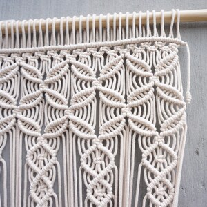 May include: A cream-colored macrame wall hanging featuring intricate knotted patterns suspended from a smooth wooden dowel. The design consists of repeating geometric shapes and vertical fringe elements created with thick cotton cord.