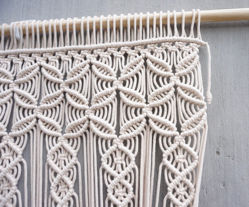 May include: A cream-colored macrame wall hanging featuring intricate knotted patterns suspended from a smooth wooden dowel. The design consists of repeating geometric shapes and vertical fringe elements created with thick cotton cord.