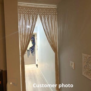 May include: A beige macrame doorway curtain featuring an intricate knotted geometric pattern at the top, transitioning into long, vertical hanging fringe strands that are gathered at the sides to create an open, arched entryway effect.