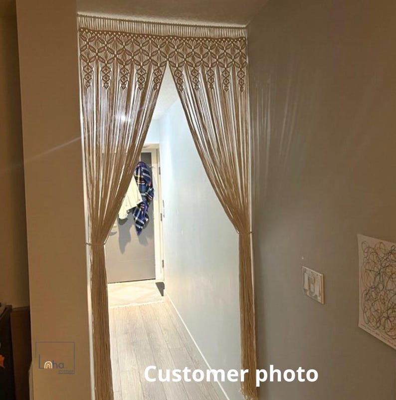May include: A beige macrame doorway curtain featuring an intricate knotted geometric pattern at the top, transitioning into long, vertical hanging fringe strands that are gathered at the sides to create an open, arched entryway effect.