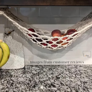 Puede incluir: Una hamaca de frutas de macram&eacute; hecha a mano llena de melocotones maduros, suspendida de una clavija de madera. Un racimo de pl&aacute;tanos cuelga de un cord&oacute;n decorativo de macram&eacute;. La encimera de la cocina es de granito.