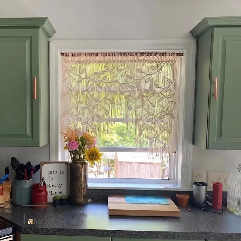 May include: A cream-colored macrame window curtain featuring a repeating leaf and vine pattern, suspended from a thin rod. The curtain has a fringe bottom edge and is positioned over a window between two sage green kitchen cabinets.