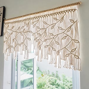 May include: A cream-colored macrame window valance hanging from a wooden rod. The textile features an intricate knotted pattern of trailing vines and leaves, with long, loose vertical fringe strands extending from the bottom edge of the design.
