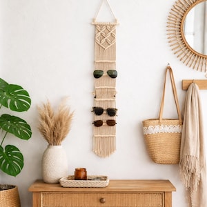 Könnte beinhalten: Ein Sonnenbrillenhalter aus beigem Makramee mit drei Sonnenbrillenpaaren. Der Halter hängt an einer weißen Wand. Ein Holztisch, ein Spiegel und andere Dekorationen sind ebenfalls zu sehen.