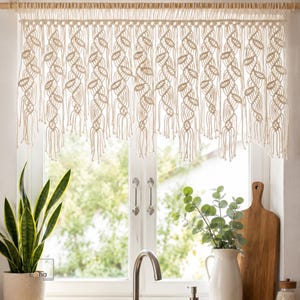 May include: A cream-coloured macrame window valance featuring a repeating leaf-like pattern created with intricate knotted cord work. The valance hangs from a wooden rod and displays a series of vertical strands ending in small, decorative fringe.
