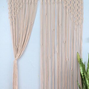 May include: A beige macrame curtain featuring intricate leaf-shaped knot patterns along the top section, transitioning into long, straight vertical cords. The curtain is suspended from a wooden rod, with one side gathered and tied together.