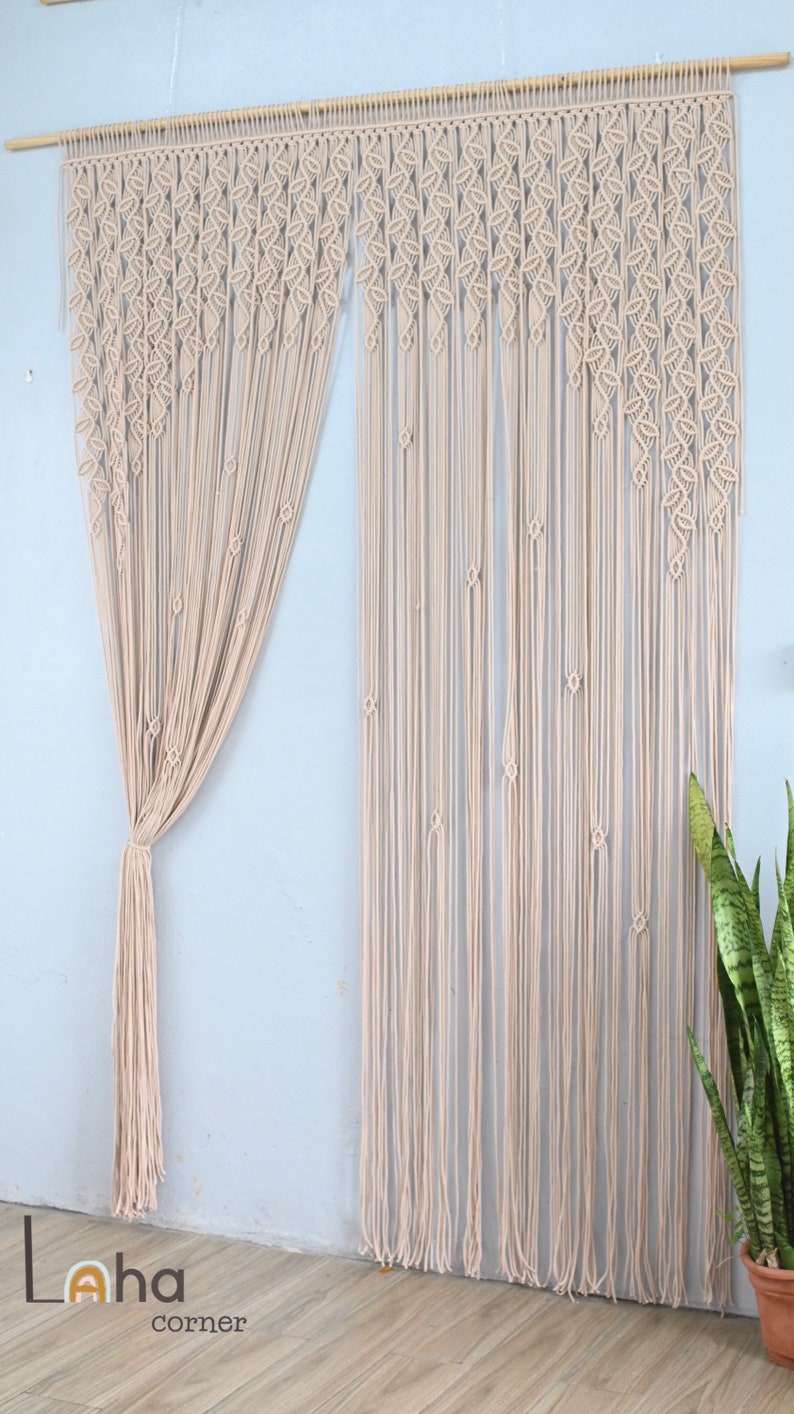 May include: A beige macrame curtain featuring intricate leaf-shaped knot patterns along the top section, transitioning into long, straight vertical cords. The curtain is suspended from a wooden rod, with one side gathered and tied together.