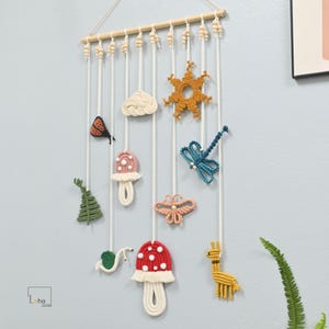 Könnte beinhalten: Ein Makramee-Wandbehang mit einem Holzstab und weißen Kordeln. Daran befestigt sind verschiedene Textilfiguren, darunter ein grüner Baum, eine Schnecke, ein rosa Pilz, eine weiße Wolke, ein roter Pilz, eine Sonne, eine Libelle, ein Schmetterling und eine gelbe Giraffe.