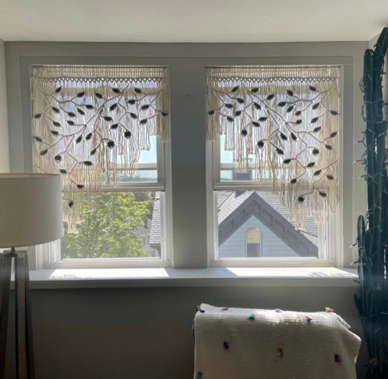 May include: Two off-white macrame window valances featuring a knotted branch and leaf pattern. Each valance has a horizontal header with vertical fringe strands hanging down, creating a decorative textile covering for the upper window panes.