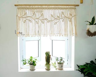 May include: A cream-colored macrame window curtain hanging from a wooden rod. The textile features a knotted leaf pattern across the top section with long, vertical fringe strands hanging down. The text Laha corner is visible on the left side.