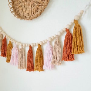 May include: A garland of wooden beads with tassels in pink, beige, and orange hues. The tassels are made of yarn and hang from the beads.