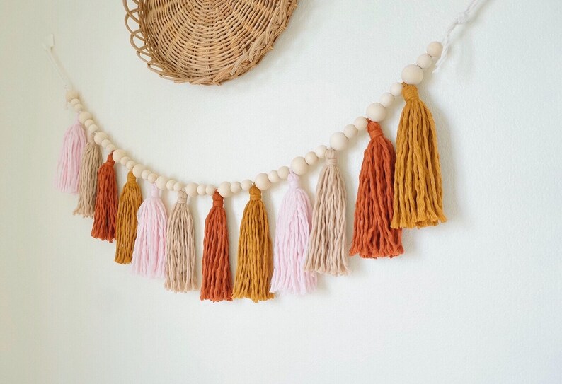 May include: A garland of wooden beads with tassels in pink, beige, and orange hues. The tassels are made of yarn and hang from the beads.