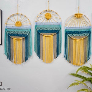 May include: Three macrame wall hangings with a blue, green, and yellow color scheme. Each hanging features a circular design with a sunburst pattern and a fringe of yarn.