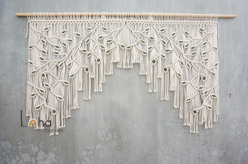 May include: A cream-colored macrame wall hanging suspended from a wooden rod, featuring an intricate knotted leaf pattern with small green accents integrated into the design. The piece has a tiered fringe bottom edge and the text Laha corner.
