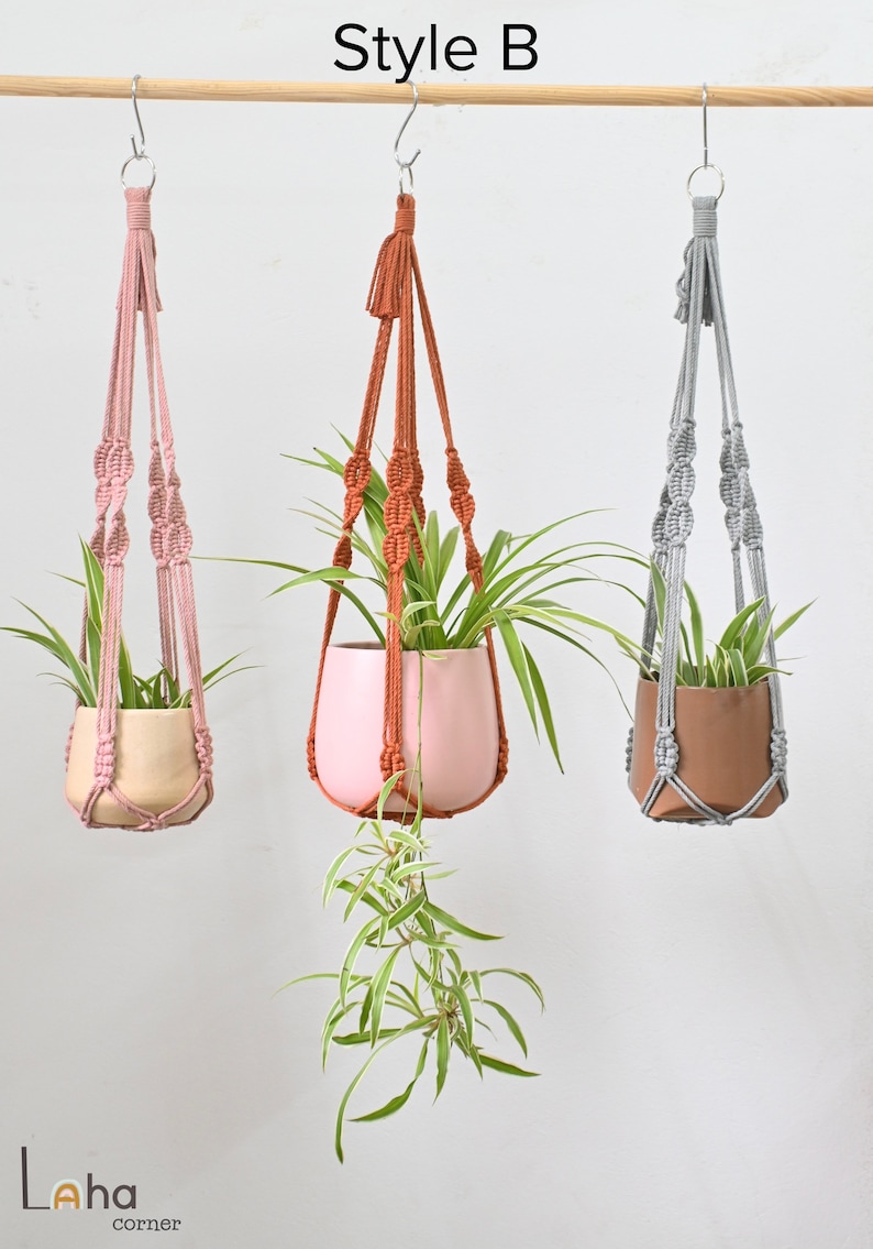 May include: Three macrame plant hangers with different colored cords and pots. The hangers are hanging from a wooden dowel rod. The pots are beige, pink, and brown. The plants are green and leafy.