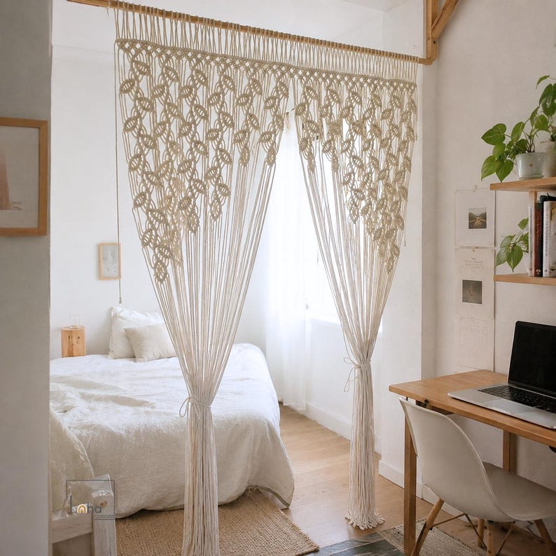 May include: A cream-colored macrame curtain featuring an intricate leaf-like knot pattern suspended from a wooden rod. The curtain is divided into two panels, each gathered and tied at the center, with long, straight fringe extending to the floor.