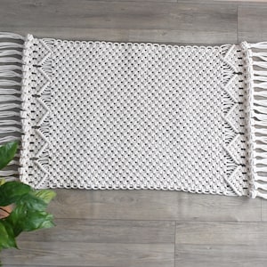 May include: A rectangular, off-white macrame rug with a geometric diamond pattern and fringed edges. The rug is made of woven cotton and is suitable for a living room or bedroom. The rug is approximately 36 inches long and 24 inches wide.