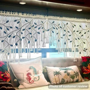 May include: A cream-colored macrame window valance featuring an intricate knotted branch and leaf pattern with hanging fringe. The design includes small blue accents integrated into the woven structure, creating a textured, decorative window covering.