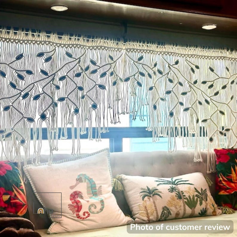 May include: A cream-colored macrame window valance featuring an intricate knotted branch and leaf pattern with hanging fringe. The design includes small blue accents integrated into the woven structure, creating a textured, decorative window covering.