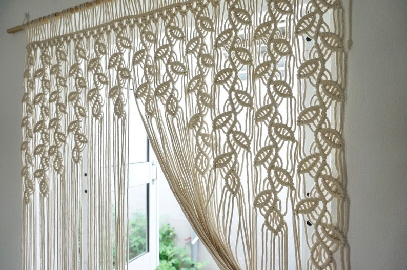 May include: An off-white macrame curtain hanging from a wooden rod, featuring a repeating leaf-like knot pattern across the top section that transitions into long, vertical, unknotted fringe strands that hang down to the bottom edge.
