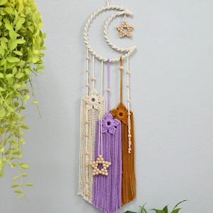 Könnte beinhalten: Makramee-Wandbehang mit Mond- und Sternenmotiv. Er besteht aus cremefarbenem, lila und braunem Garn mit Akzenten aus Holzperlen. Das Design beinhaltet ein sternförmiges Element aus Holzperlen.