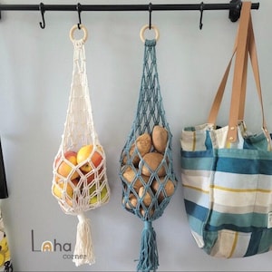 May include: Two macrame hanging produce bags with wooden rings, one cream and one blue, filled with fresh fruit and potatoes respectively, hanging from a black metal rack alongside a blue, white, and yellow striped canvas tote bag with leather straps.