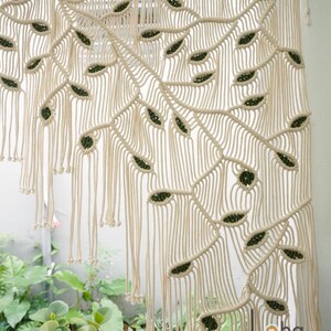 May include: A macrame wall hanging featuring off-white cotton cord knotted into a pattern of large, sweeping branches with dark green leaf-shaped accents. The piece hangs from a wooden rod and includes the text Laha corner in the bottom right.