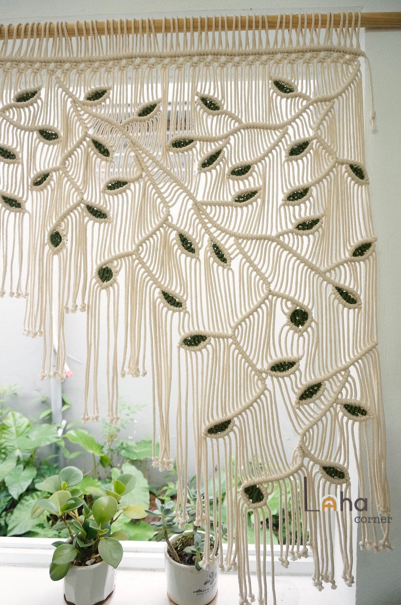 May include: A macrame wall hanging featuring off-white cotton cord knotted into a pattern of large, sweeping branches with dark green leaf-shaped accents. The piece hangs from a wooden rod and includes the text Laha corner in the bottom right.