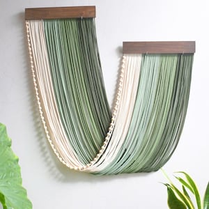 May include: A wall hanging featuring two dark brown wooden bars from which long, draped strands of cream and sage green cord are suspended. A single string of small, round, light-coloured wooden beads follows the curve of the draped cord.