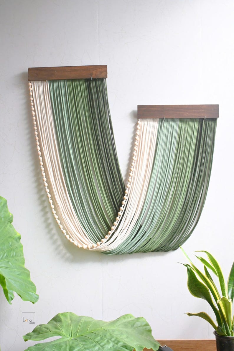 May include: A wall hanging featuring two dark brown wooden bars from which long, draped strands of cream and sage green cord are suspended. A single string of small, round, light-coloured wooden beads follows the curve of the draped cord.