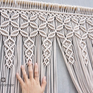 May include: An off-white macrame wall hanging featuring a repeating geometric pattern of interconnected circular knots suspended from a wooden dowel, with long vertical fringe strands extending downward and the text Laha corner visible.