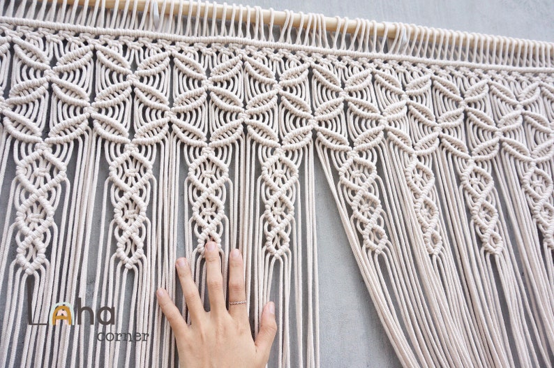 May include: An off-white macrame wall hanging featuring a repeating geometric pattern of interconnected circular knots suspended from a wooden dowel, with long vertical fringe strands extending downward and the text Laha corner visible.