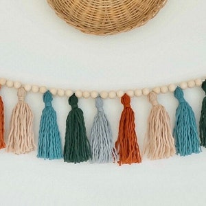 May include: A garland of wooden beads with colorful yarn tassels in shades of orange, blue, green, and beige. The garland is hanging on a white wall.