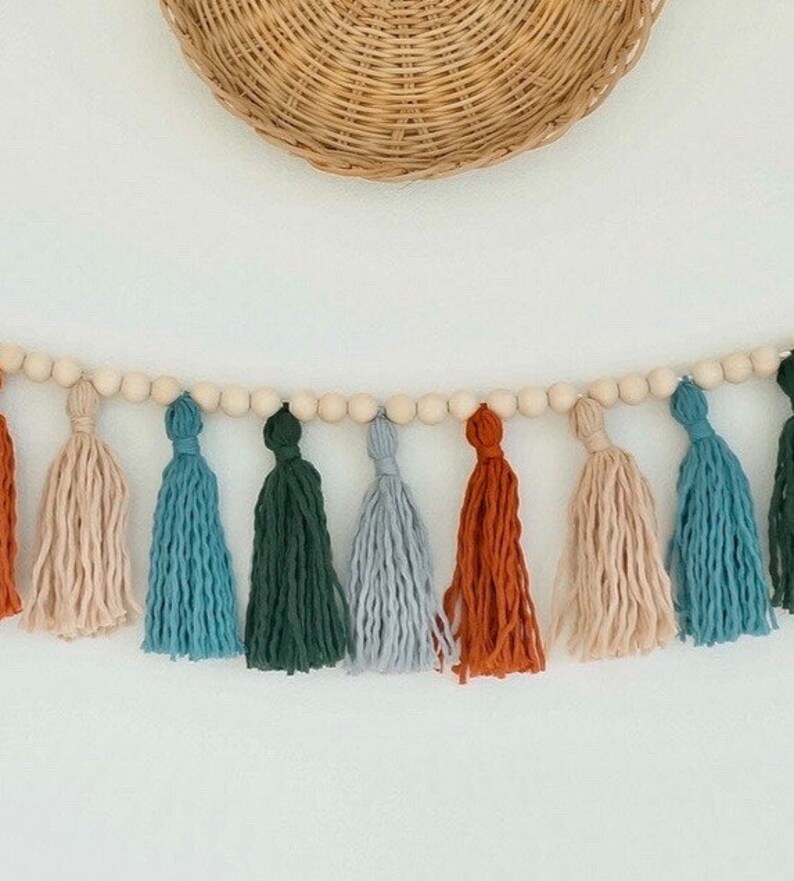 May include: A garland of wooden beads with colorful yarn tassels in shades of orange, blue, green, and beige. The garland is hanging on a white wall.