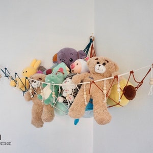 May include: A white string hammock with wooden beads hangs in a corner of a room. It is filled with stuffed animals, including a brown teddy bear, a green dinosaur, a purple elephant, and a yellow bunny.  There are rainbow wall decorations on either side of the hammock.