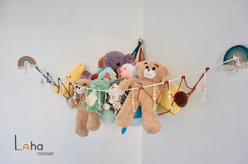 May include: A white string hammock with wooden beads hangs in a corner of a room. It is filled with stuffed animals, including a brown teddy bear, a green dinosaur, a purple elephant, and a yellow bunny.  There are rainbow wall decorations on either side of the hammock.
