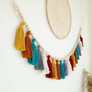 May include: A colorful tassel garland with wooden beads. The garland features tassels in shades of yellow, orange, teal, gray, and red. The garland is strung on a white cord and is perfect for adding a touch of bohemian style to any room.