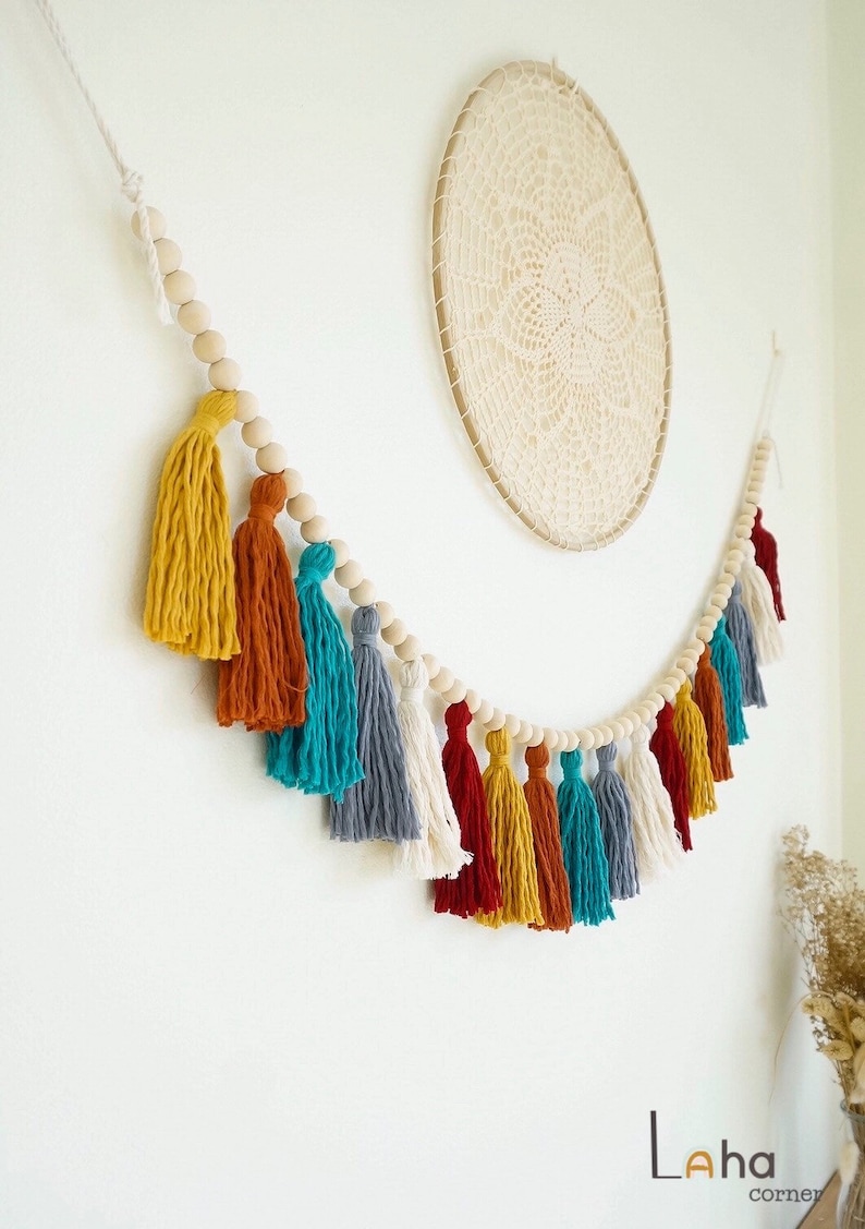 May include: A colorful tassel garland with wooden beads. The garland features tassels in shades of yellow, orange, teal, gray, and red. The garland is strung on a white cord and is perfect for adding a touch of bohemian style to any room.