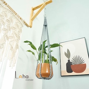 Puede incluir: Una maceta colgante con un colgador de macramé gris. La maceta de color terracota contiene una planta verde. La maceta está suspendida de un soporte de madera fijado a la pared. También se ven una impresión artística enmarcada y una cortina de macramé.