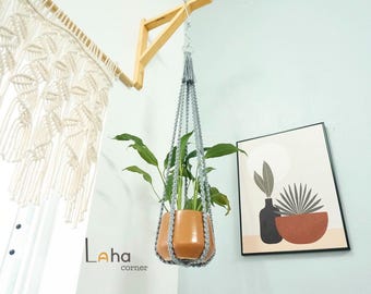 May include: A hanging plant pot with a gray macrame hanger. The terracotta-colored pot holds a green plant. The pot is suspended from a wooden bracket attached to the wall. A framed art print and a macrame curtain are also visible.