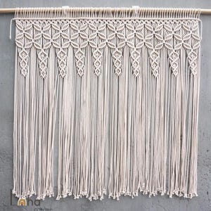 May include: A cream-colored macrame wall hanging suspended from a wooden rod. The piece features a repeating geometric pattern of intricate knots across the top section, transitioning into long, vertical, unknotted fringe strands at the bottom.