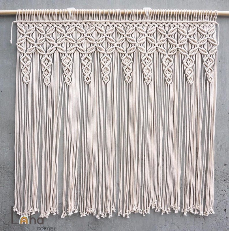 May include: A cream-colored macrame wall hanging suspended from a wooden rod. The piece features a repeating geometric pattern of intricate knots across the top section, transitioning into long, vertical, unknotted fringe strands at the bottom.