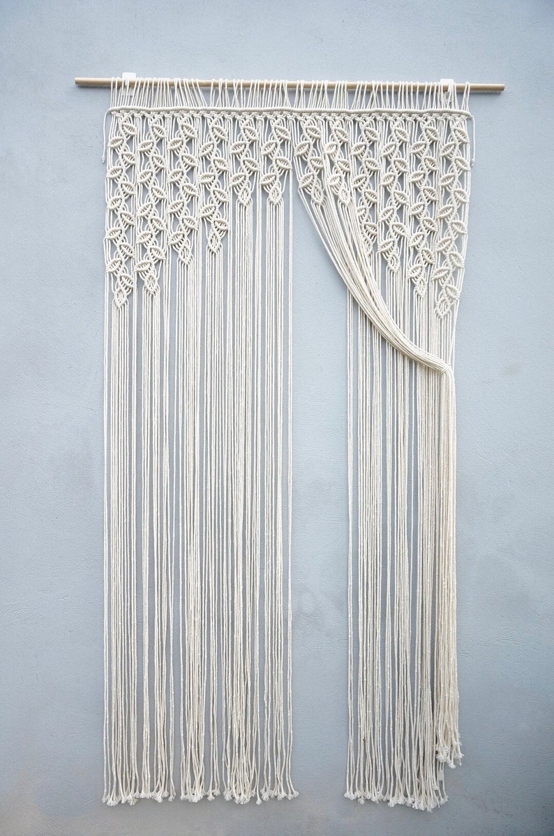 May include: A cream-colored macrame wall hanging suspended from a wooden rod. The upper section features a dense pattern of knotted leaf-like shapes, while the lower portion consists of long, vertical, unknotted fringe strands hanging straight down.