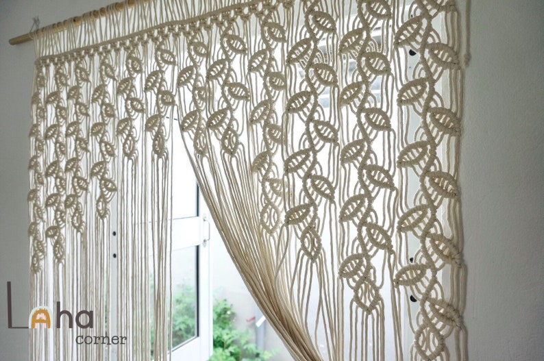 May include: An off-white macrame curtain hanging from a wooden rod, featuring a repeating leaf-like knot pattern and long vertical fringe strands. The lower left corner displays the text Laha corner in a dark gray font with a small orange icon.