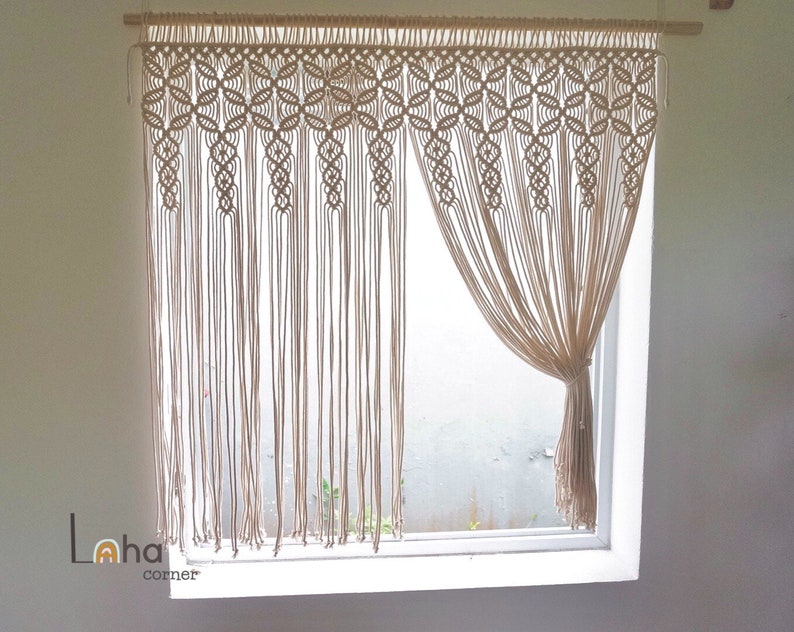 May include: A beige macrame curtain with a geometric pattern hangs in a window. The curtain is made of natural fibers and has a fringed bottom.