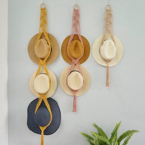 Peut inclure: Trois suspensions murales en macramé faites de corde jaune moutarde, rose poudré et crème. La suspension jaune porte trois chapeaux, la rose porte deux chapeaux et la crème porte un chapeau. Chaque suspension possède un anneau en bois au sommet.