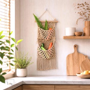May include: A beige macrame wall hanging with two tiered storage pockets suspended from a wooden dowel. The woven cord structure features an open diamond pattern and a bottom fringe. The pockets contain green fern leaves, a carrot, and cucumbers.