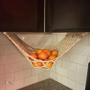 May include: A macrame fruit hammock filled with oranges, suspended under a dark cabinet. The hammock is made of beige rope and hangs against a white tiled backsplash. The oranges are bright orange.
